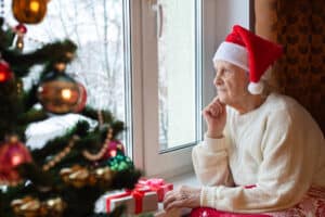 Elderly man wearing a Santa hat sits by a decorated Christmas tree.