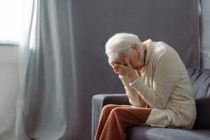 Older woman sitting on gray couch with head leaning on hands.