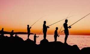 Silhouettes of people fishing by a body of water at sunset.