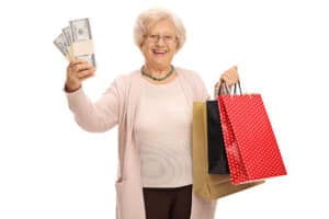 Elderly woman holding stacks of money and shopping bags.